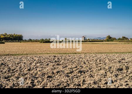 Terreno agricolo arato che mostra terreno fertile pronto per la semina, con un campo raccolto, alberi distanti, edifici e un silo di grano sotto un bl trasparente Foto Stock