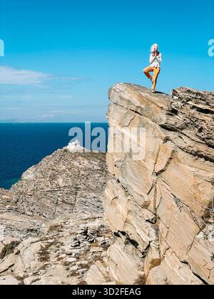 Una donna si erge nell'albero dello yoga posato su una cima rocciosa di fronte al faro nella riserva naturale del Parco di Paros, in Grecia. Foto Stock