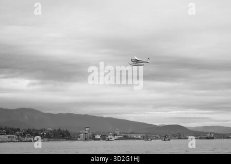 Un idrovolante vola sopra Burrard Inlet in questa scena in bianco e nero che mostra l'industria portuale, le imbarcazioni e le sagome di montagne lontane. Foto Stock