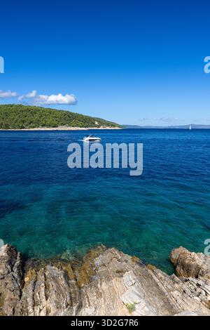Un tranquillo paesaggio marino con una barca a vela solitaria che scivola su acque blu profonde, un primo piano roccioso e un'isola verdeggiante in lontananza sotto un luminoso e chiaro sk Foto Stock