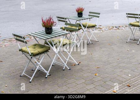 Una fila di tavoli e sedie da caffè all'aperto vuoti e pieghevoli, verdi e bianchi, con cuscini d'oliva e piccole piante di erica su un marciapiede lastricato. Foto Stock