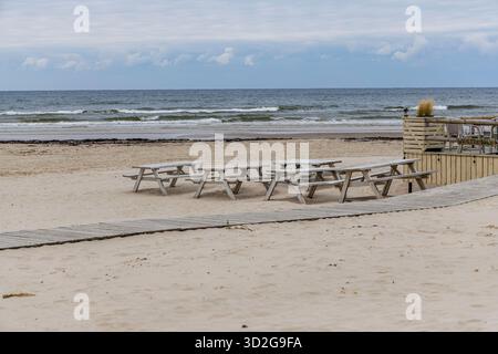Diverse file di tavoli da picnic in legno seduti su un'ampia e vuota spiaggia di sabbia, con una passerella di legno che conduce ad una terrazza caffè vicino al mare. Foto Stock