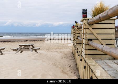 Vista prospettica di una terrazza in legno con lanterne decorative e un tavolo da picnic parzialmente coperto da sabbia soffiata dal vento sulla spiaggia. Foto Stock