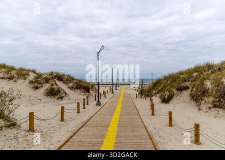 Vista diretta e centrale di una passerella di legno con una striscia gialla brillante che corre lungo il centro, fiancheggiata da dune di sabbia, che conduce verso il mare nel Foto Stock