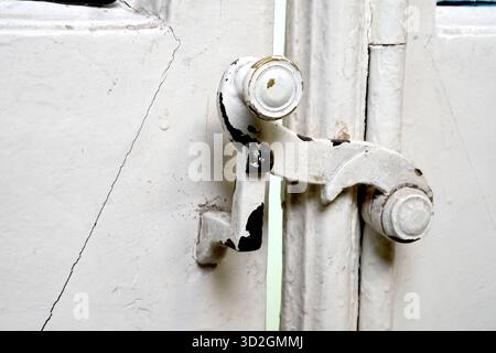 Un'immagine macro di un vecchio dispositivo di chiusura in metallo bianco su una porta o una finestra con vernice spaccata e staccata, su uno sfondo bianco con una spaccatura evidente. Foto Stock