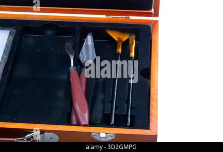 Primo piano di una cassa d'arte aperta in legno rivestita di materiale nero, contenente coltelli palette con manico rosso e spazzole a ventaglio per la pittura. Foto Stock