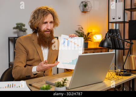 Un uomo d'affari di Redhead che lavora su un laptop alla scrivania parla di videochiamata di comunicazione online con il dipendente, il capo mostra analisi grafiche. I servizi di supporto del ragazzo caucasico guardano la webcam del computer Foto Stock