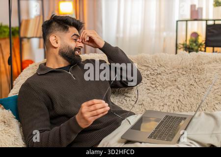 L'uomo indiano a casa strofina gli occhi con espressioni stanche mentre usa il laptop, affaticato dall'esposizione a lungo schermo. Il ragazzo arabo sul divano mostra disagio e affaticamento degli occhi, lottando contro lo stress da sovraccarico digitale Foto Stock