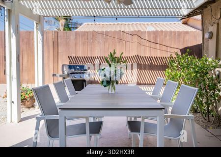 Tavolo all'aperto nel patio sul retro, sedie, grigliate, bouquet floreale con ortensie blu e gemme di giglio in vaso di vetro. Recinzione e pergolato per la privacy. Foto Stock