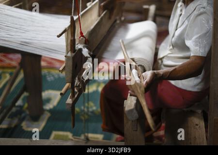 Donna asiatica concentrata che tessera con tradizionale abilità artigianale su telaio in legno. Un artigiano lavora con la navetta e il filo bianco per creare un bellissimo textil Foto Stock