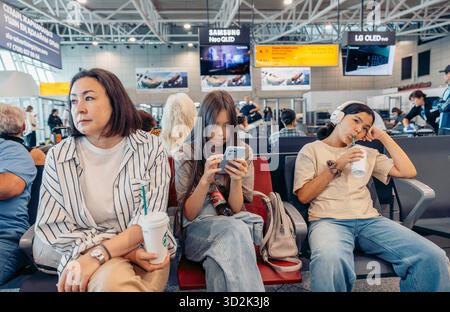 ALMATY, KAZAKISTAN - 18 AGOSTO 2025: Tre giovani donne siedono in un'area d'attesa dell'aeroporto. Sono impegnati in una conversazione Foto Stock