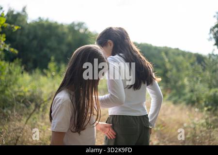 Due ragazze nella foresta che si piegano e cercano qualcosa per terra. Erba dorata sfocata e alberi sullo sfondo, luce naturale soffusa, Co Foto Stock