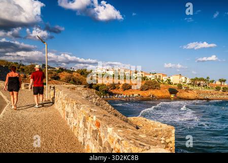 Panormos, Creta, Grecia - 2 settembre 2025: Turisti che camminano su una passeggiata in pietra sul mare con il paesaggio costiero panoramico e il cielo estivo blu. Foto Stock