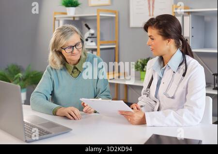Paziente anziano seduto alla scrivania in ufficio medico parlare con giovane medico donna. Foto Stock