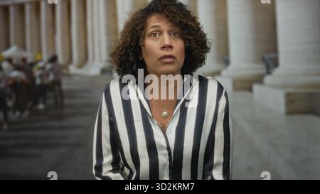 Donna con capelli ricci e collana con medaglioni che guarda in alto le colonne di costruzione sulla strada della città vecchia; stupore curiosità. Foto Stock