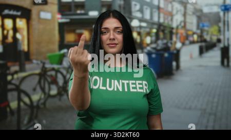 Una donna in camicia verde volontaria in piedi per strada fa un gesto scortese, mostrando il dito medio, con sfondo urbano sfocato. Foto Stock