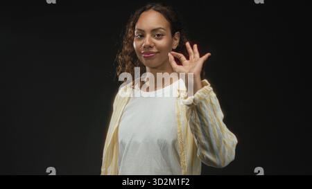 La giovane donna afroamericana punta il dito con il viso in avanti indossando t shirt bianca e camicia a righe gialle in studio nero; sicurezza e facilità. Foto Stock
