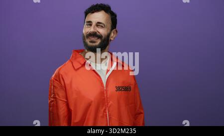 Uomo ispanico in una tuta di prigione arancione sorridente con fiducia su un vibrante sfondo viola. Foto Stock