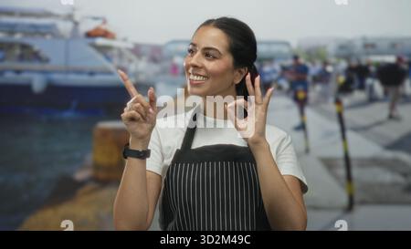 La donna che indossa un grembiule in tessuto nero sorride mentre fa un cartello con la mano destra e punta il dito con la sinistra al molo del porto; servizio cortese per l'approvazione. Foto Stock