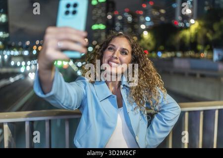 Giovane donna con i capelli ricci sorride felicemente mentre scatta un selfie sul suo cellulare, in piedi su un ponte che si affaccia sui semafori della città e sul traffico Foto Stock