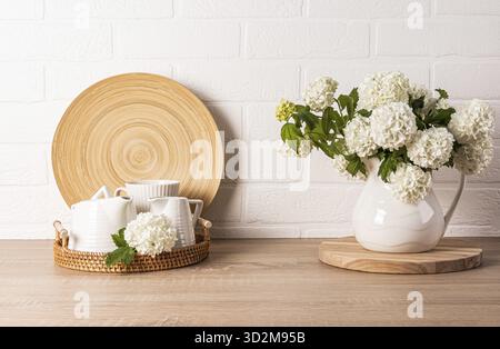 Set da tè in ceramica bianca su vassoio rotondo in vimini sul piano della cucina. fiori decorativi nella caraffa. Sfondo della cucina in colori chiari. Vista frontale Foto Stock