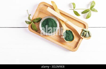 Un vasetto di vetro di polvere di spirulina verde biologica e due cucchiai di legno riempiti di polvere e capsule su un vassoio di legno Vista dall'alto. Disintossicazione Foto Stock