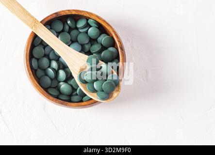 Primo piano di compresse verdi di alga spirulina benefica su un cucchiaio di legno in una ciotola di legno riempita di compresse. Vista dall'alto. Disintossicazione Foto Stock