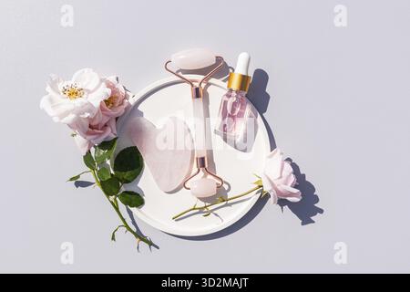Una bottiglia trasparente con un prodotto cosmetico con siero o olio di rosa giace in un piatto con petali di rosa, un massaggiatore al quarzo, un raschietto gua Sha. Vista dall'alto Foto Stock