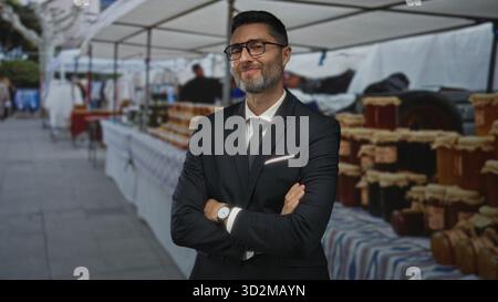 Uomo in tuta e cravatta con occhiali e barba, braccia incrociate che mostrano le mani alle affollate bancarelle del mercato di strada con vasi e tavoli; tranquillità. Foto Stock