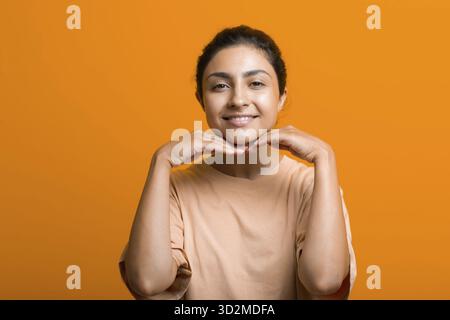 Primo piano ritratto della giovane donna indiana americana toccando pelle del viso le sue mani e facendo auto yoga viso ginnastica massaggio arancione sfondo Foto Stock