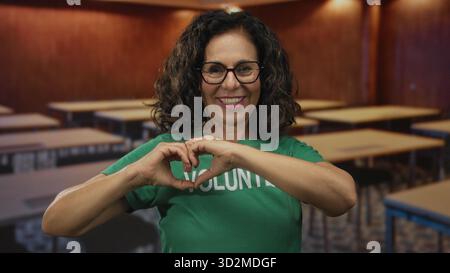 Donna volontaria che fa un gesto di cuore in classe mentre sorride con una camicia verde all'interno con scrivanie, rappresentando il sostegno della comunità e po Foto Stock