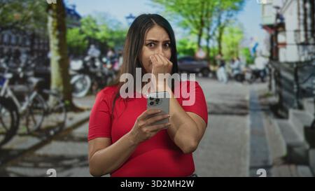 Donna che tiene lo smartphone pizzicando il naso sulla strada accanto alle biciclette parcheggiate e ai gradini, indossa una camicia rossa e sembra scontenta; disgusto per la preoccupazione. Foto Stock