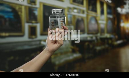 Tenendo a mano un vaso di vetro con dollari americani all'interno di un museo, illustrando temi finanziari con dipinti sfocati sullo sfondo elegante Foto Stock