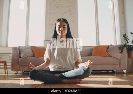Donna asiatica che fa yoga e zen come meditazione lotus posa in casual abbigliamento in appartamento soggiorno interno con luce naturale del sole Foto Stock
