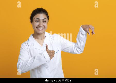 Ritratto della donna medica indiana in abito medico bianco che tiene le dita modello vaccino covid-19 o altre cose mediche isolate su sfondo giallo Foto Stock