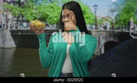 La donna latina tiene una ciotola di vetro di patatine fritte e dà un gesto in giro per strada; approvazione allegria. Foto Stock