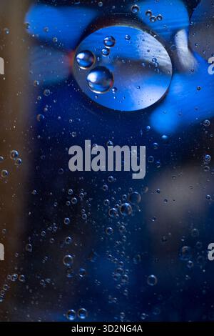 Bolle d'olio di varie dimensioni che galleggiano sulla superficie dell'acqua su uno sfondo blu sfocato. Foto Stock