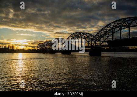 Iconico ponte ferroviario ad arco in acciaio sul fiume Daugava a riga al tramonto, che mostra un cielo spettacolare, riflessi sull'acqua e silhouette di trasposizione storica Foto Stock