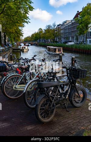 Biciclette parcheggiate lungo il canale di Amsterdam con barche e ponti Foto Stock