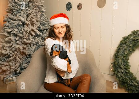 una bellissima ragazza riccia con un maglione bianco dai capelli scuri si siede su una poltrona e tiene in braccio un dachshund nero. Ragazza con un cappello di Babbo Natale. Felice nuova Yea Foto Stock