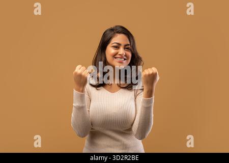 Concetto di vincita. Una donna araba gioiosa che festeggia la vittoria, scuotendo i pugni arroccati sullo sfondo beige dello studio Foto Stock