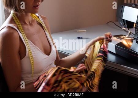 Bella giovane donna seduta di fronte a una macchina da cucire che regge il tessuto liscio di raso, preparandosi per il cucito. Crea stilista di moda in corso. Foto Stock