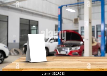 Carta bianca vuota in piedi su un tavolo di legno nel garage di servizio auto con sfondo sfocato delle auto in manutenzione, ideale per il mockup, la pubblicità Foto Stock