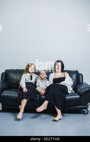 Una madre e i suoi due figli si godono un momento di connessione su un divano in pelle nera. Condividono sorrisi e risate, mostrando la loro stretta famiglia bon Foto Stock