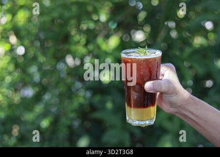 Rinfrescante caffè a strati ghiacciato in vetro tenuto a mano all'aperto. fresca bevanda estiva con guarnizione alla menta gustata contro una splendida natura verdeggiante Foto Stock