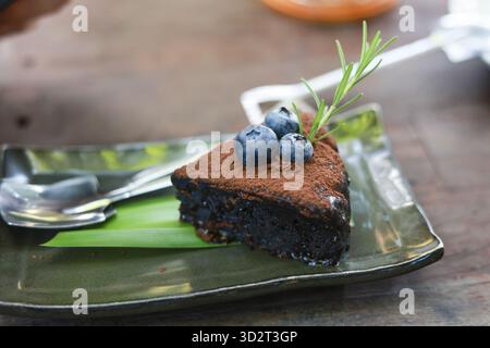 Deliziosa fetta di torta al cioccolato fondente su un piatto verde. Dessert decadente ricoperto di cacao in polvere, mirtillo fresco e rametto di rosmarino per buongustai Foto Stock