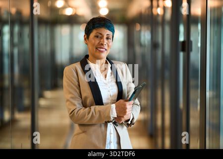 Donna d'affari matura che sorride con fiducia nel corridoio dell'ufficio in posa Foto Stock