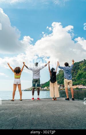 Immagine verticale di un gruppo eterogeneo di giovani amici in piedi su un molo, tenendosi per mano con le braccia alzate per celebrare l'unità e il successo. Foto Stock