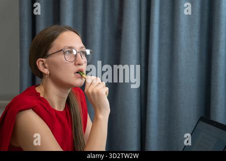 Una donna d'affari che indossa gli occhiali è seduta alla scrivania con un computer portatile e tiene la penna vicino alle labbra, pensando al progetto e lavorando in remoto da casa Foto Stock