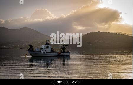 Una piccola barca da pesca e un pescatore che tirano le reti all'alba con il sole autunnale che si infrangono tra le nuvole. Foto Stock